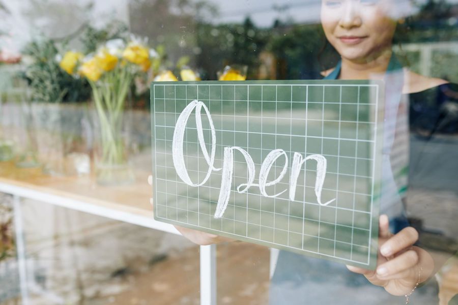 Woman smiling and holding an open sign outside a colorful flower shop filled with blooming plants.